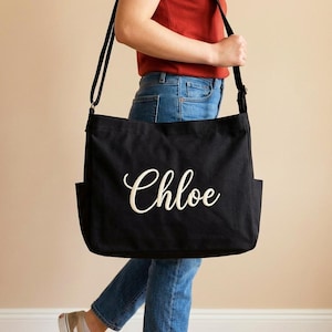 May include: Black canvas tote bag with the name "Chloe" in white script. The bag has a long shoulder strap and two exterior pockets. A casual and practical accessory.