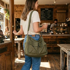 May include: A woman in a cafe, carrying a large olive green canvas shoulder bag with multiple pockets. The bag has a long adjustable strap. The cafe has wooden tables and chairs, and a sign in the window reads "OPEN".
