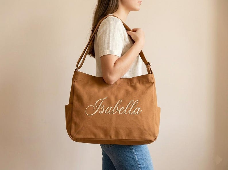 May include: A brown canvas tote bag with the name "Isabella" embroidered in a cursive font. The bag has two exterior pockets and a long shoulder strap. The bag is carried over the shoulder.