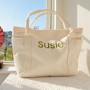 May include: Cream-colored canvas tote bag with two exterior pockets and two handles. The word "Susie" is embroidered in green on the front of the bag. The bag is sitting on a white lace tablecloth.
