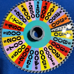 May include: A colourful wheel game with a variety of dollar amounts, including $150, $200, $300, $400, $500, $700, $800, $900, $1000, $2000, $4000, and $5000. The wheel is divided into sections of different colours, with each section containing a dollar amount. The wheel is made of cardboard and has a metal centre.