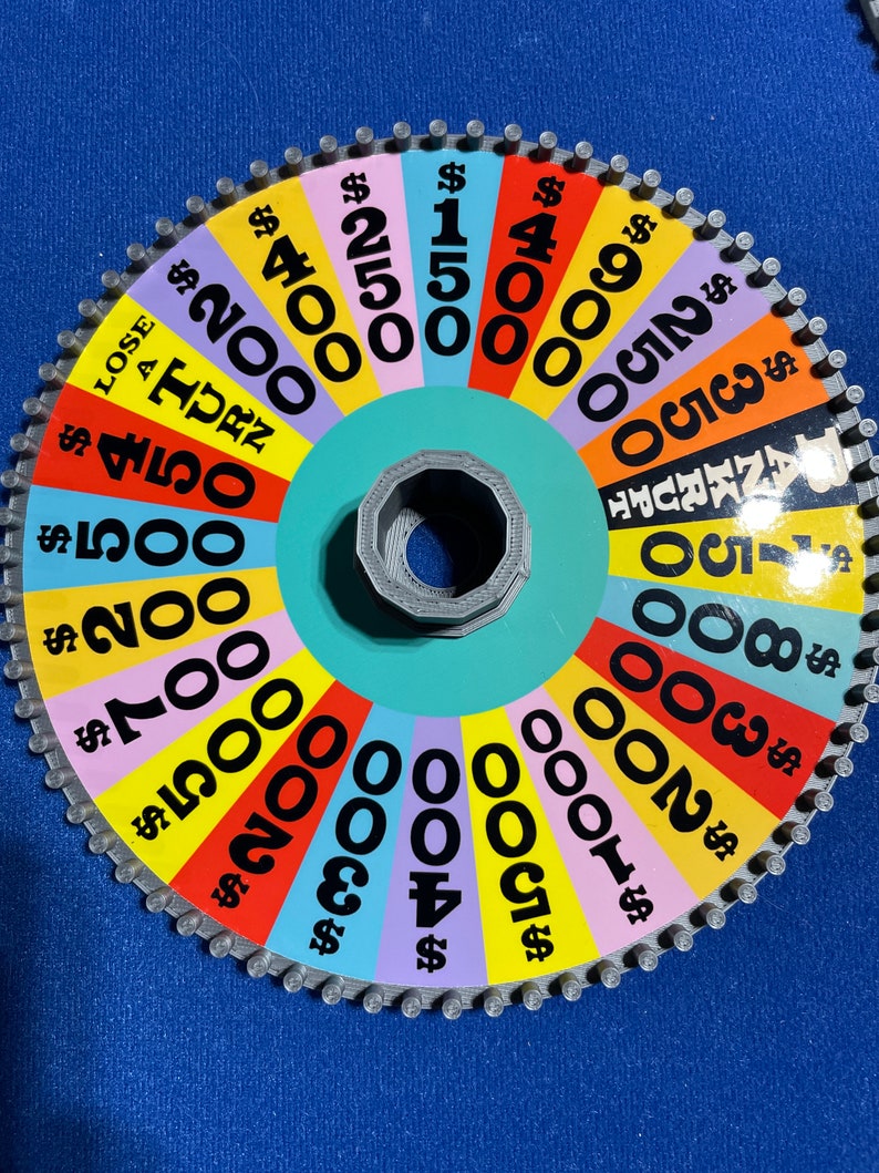 May include: A colourful wheel game with a variety of dollar amounts, including $150, $200, $300, $400, $500, $700, $800, $900, $1000, $2000, $4000, and $5000. The wheel is divided into sections of different colours, with each section containing a dollar amount. The wheel is made of cardboard and has a metal centre.