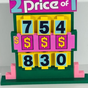 Puede incluir: Una colorida exhibición de precios de juguetes con el texto "for the price of 2 for 1". La pantalla muestra los números 754 y 830, junto con símbolos de dólar. La pantalla está hecha de plástico verde, rosa, amarillo y azul.