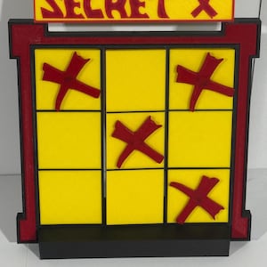 Puede incluir: Un tablero de tres en raya amarillo y negro con X rojas. El tablero tiene un marco rojo y negro con un letrero que dice "SECRET "X"".