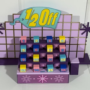 Puede incluir: Un expositor morado y dorado con el texto "1/2 Off" en la parte superior. Presenta dieciséis cajas numeradas en varios colores, dispuestas en una cuadrícula. El expositor tiene detalles en forma de estrella.