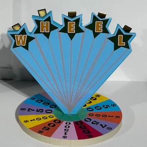 Puede incluir: Un juego de la rueda de la fortuna azul y blanco con una rueda giratoria y un puntero en forma de estrella con la palabra "WHEEL" en él.