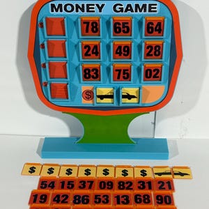 Puede incluir: Un tablero de juego azul y naranja con las palabras "MONEY GAME" impresas en él. El tablero tiene 12 casillas con números impresos en ellas. También hay 21 casillas naranjas con números impresos en ellas. El tablero de juego está sobre una base azul.