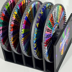 Puede incluir: Un conjunto de ruedas de juego coloridas con segmentos que muestran valores monetarios y la palabra "LOSE". Las ruedas están alojadas en un soporte negro, con un gráfico en el lateral. Las ruedas son de varios colores.