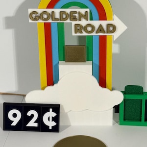 Puede incluir: Un letrero colorido impreso en 3D con las palabras "GOLDEN ROAD" y una flecha. El letrero presenta un diseño de arcoíris con una nube blanca y un pequeño objeto cuadrado. Un letrero negro muestra "92¢".