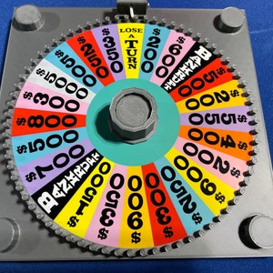 May include: A colourful wheel of fortune game with a spinning wheel and a grey base. The wheel has different dollar amounts and the phrase "Lose a Turn".
