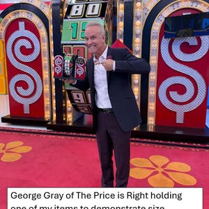 May include: George Gray, host of the game show "The Price is Right", holds a small item in front of a large spinning wheel with dollar signs on either side. The wheel has numbers from 1 to 100.