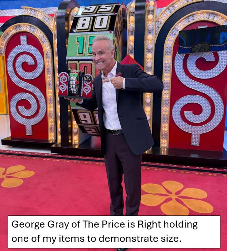 May include: George Gray, host of the game show "The Price is Right", holds a small item in front of a large spinning wheel with dollar signs on either side. The wheel has numbers from 1 to 100.