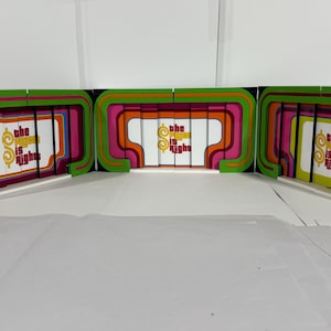 Puede incluir: Un conjunto de tres telones de fondo de cartón de colores para un plató de programa de juegos. Cada telón de fondo presenta un diseño abstracto curvo en tonos de verde, naranja, rosa y morado. El telón de fondo central tiene las palabras "The Price is Right" en letras amarillas.