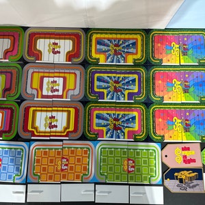 Puede incluir: Un conjunto de piezas de tablero de juego para el programa de juegos "The Price is Right". Las piezas son coloridas y presentan el logotipo del programa. Hay varios diseños diferentes, que incluyen un escenario, una etiqueta de precio y una cuadrícula.