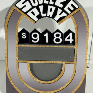 Puede incluir: Un letrero decorativo gris y dorado con las palabras "SQUEEZE PLAY" en letras blancas. El letrero muestra el número "9184" en números blancos. Tiene una ranura rectangular y un recorte semicircular.