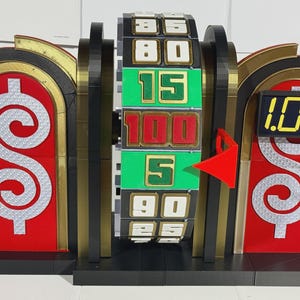 Puede incluir: Una colorida rueda de programa de juegos con una flecha roja que apunta a un número. La rueda tiene varios números en diferentes colores. La pantalla muestra "1.00". El diseño incluye elementos rojos, negros, dorados y plateados, con motivos de signo de dólar.