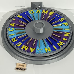 Puede incluir: Una rueda de juego gris y azul con las palabras "America's Game Spin & Win" en amarillo. La rueda tiene radios dorados y una pequeña tarjeta "Wheel of Fortune Million".