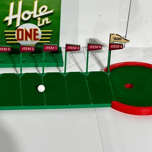 Puede incluir: Un juego de minigolf con una superficie de juego de fieltro verde y un agujero rojo y verde. El juego incluye seis banderas con números y las palabras "ITEM 1" a "ITEM 6". El juego también incluye una bola de golf blanca y un cartel que dice "Hole in ONE".