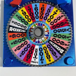 Puede incluir: Una colorida rueda de la fortuna con un centro gris y un borde exterior plateado. La rueda tiene diferentes cantidades de dólares, incluyendo $ 500, $ 600, $ 700, $ 800, $ 900 y $ 1000. La rueda también tiene las frases "Quiebra" y "Pierde un turno".