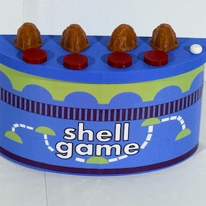 Puede incluir: Un juego de "shell game" azul y verde con cuatro piezas en forma de concha marrones, cuatro discos rojos y un disco blanco. El juguete tiene las palabras "shell game" en blanco en la parte delantera.