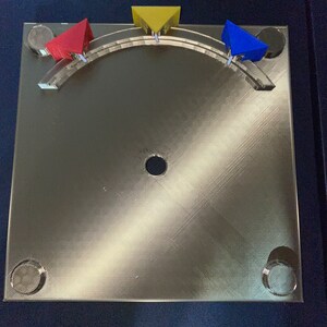 May include: A gold-coloured metal base with a circular hole in the centre. Three triangular pieces are attached to the base, one red, one yellow, and one blue.