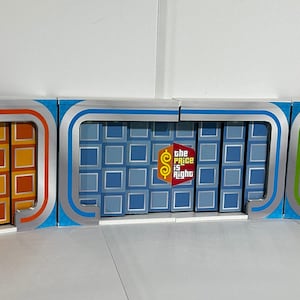 Puede incluir: Tres puertas de un programa de juegos de The Price is Right. Las puertas son de madera y tienen un patrón de cuadrícula. Las puertas están pintadas de naranja, azul y verde. Cada puerta tiene un marco plateado y una manija plateada. Las puertas están cerradas.