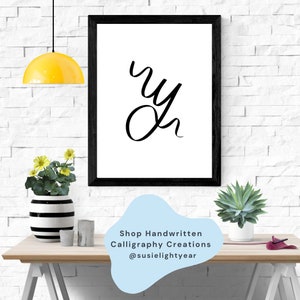 May include: Black and white framed print with the word "oh" written in cursive calligraphy. The print is on a wooden table with a yellow lamp, a black and white striped vase with yellow flowers, and a green succulent plant in a white pot.