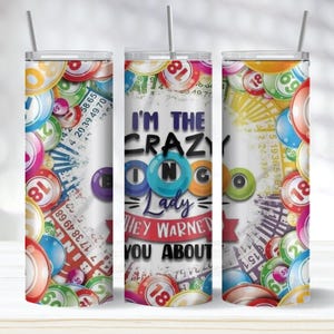 May include: A white tumbler with a straw, decorated with colorful bingo balls and cards. The text "I'M THE CRAZY BINGO Lady HEY WARNET YOU ABOUT" is displayed on the tumbler. A fun gift for bingo enthusiasts.