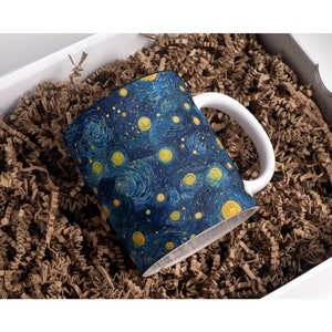 May include: A white ceramic mug featuring a blue and yellow starry night design. The mug is nestled in a box filled with brown packing paper.