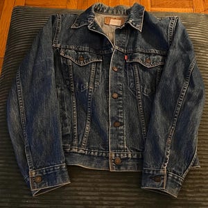 May include: A classic blue denim jacket with button closures, chest pockets, and a collar. The jacket has a worn appearance and a red tag on the chest pocket. The jacket is displayed on a green cushion.