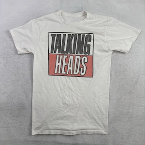 May include: White short-sleeve t-shirt featuring the band name "TALKING HEADS" in a black and red rectangular design. The band's name is printed in a bold, blocky font. The shirt is made of a soft, comfortable material.