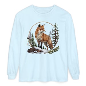 May include: A light blue long-sleeve t-shirt featuring a central circular graphic of a red fox standing on a snow-covered log. The scene includes evergreen trees, pine cones, and red berries, all framed within a thin, circular wooden-colored border.