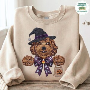 Puede incluir: Una sudadera beige de cuello redondo con un gráfico impreso de un perro doodle marrón que lleva un sombrero de bruja negro con una pequeña araña. El perro tiene una pajarita morada con un patrón de fantasmas y un caramelo de maíz central.