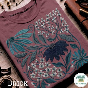 Könnte beinhalten: Ziegelrotes T-Shirt mit einem Blumendesign in Türkis, Marineblau und Weiß. Das Design umfasst große Blumen, Blätter und kleine Blüten. Das Wort "BRICK" ist unten links auf dem T-Shirt aufgedruckt.