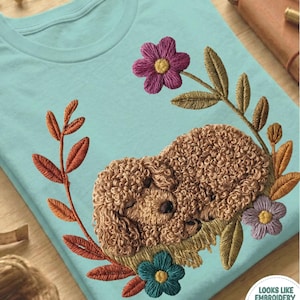Può includere: Una maglietta azzurra con un disegno ricamato di un cane marrone addormentato circondato da fiori e foglie colorate. Il testo "CHALKY MINT" è stampato in basso sulla maglietta.