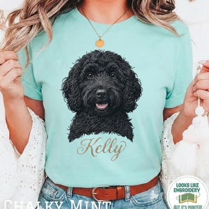 May include: A mint green t-shirt featuring a black dog portrait with the name "Kelly" in gold script. The shirt also has the words "Chalky Mint" printed at the bottom. The design gives the impression of embroidery.