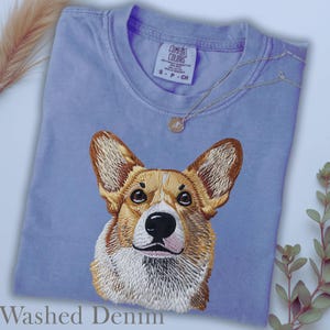 K&ouml;nnte beinhalten: Hellblaues T-Shirt mit gesticktem Corgi-Hund-Design. Das Shirt hat einen Rundhalsausschnitt und den Text "Washed Denim" unten. Eine silberne Halskette mit kleinem Anh&auml;nger liegt auf dem Shirt.