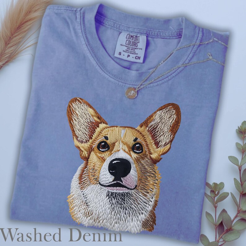 K&ouml;nnte beinhalten: Hellblaues T-Shirt mit gesticktem Corgi-Hund-Design. Das Shirt hat einen Rundhalsausschnitt und den Text "Washed Denim" unten. Eine silberne Halskette mit kleinem Anh&auml;nger liegt auf dem Shirt.