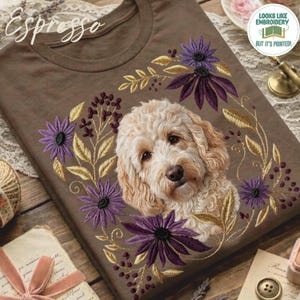May include: A brown t-shirt featuring an embroidered portrait of a curly-haired golden dog surrounded by a wreath of purple flowers and gold leaves. The word Espresso is written in a cursive script at the top left of the brown fabric.