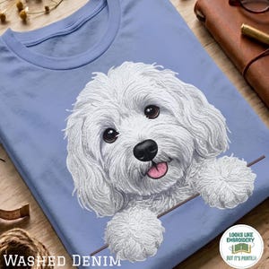 以下が含まれることがあります： 明るい青色のTシャツで、暗い目とピンクの舌を持つふわふわした白い犬のグラフィックイラストが描かれています。犬は水平な線の向こうから顔を覗かせているように見えます。隅にある小さな円形のロゴには、Looks Like Embroideryというテキストが含まれています。