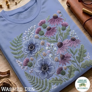 May include: A light blue t-shirt featuring a detailed floral embroidery design. The pattern includes purple coneflowers, blue anemones, green fern leaves, and small white blossoms. A circular logo in the corner reads LOOKS LIKE EMBROIDERY BUT IT'S PRINTED.