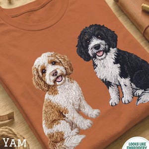 May include: A terracotta-coloured t-shirt with embroidered-style images of two dogs. One dog is brown and white, the other is black and white. The shirt has the word "YAM" printed on it, and a logo that says "Looks Like Embroidery But It's Printed!"