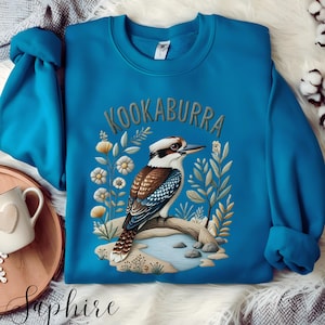 Könnte beinhalten: Ein türkisfarbenes Sweatshirt mit einer detaillierten Illustration eines Kookaburra-Vogels, der auf einem Ast sitzt, umgeben von Blumen und Blättern. Das Wort "KOOKABURRA" ist über dem Vogelbogen angeordnet. Das Sweatshirt hat einen Rundhalsausschnitt und lange Ärmel.