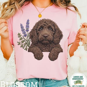 May include: Pink t-shirt featuring a detailed illustration of a brown dog with curly fur, accompanied by lavender sprigs. The word "BLOSSOM" is printed at the bottom. The shirt also has a small circular logo that says "LOOKS LIKE EMBROIDERY BUT IT'S PRINTED!"