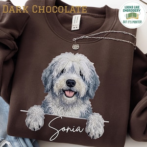 Könnte beinhalten: Ein dunkel schokoladenbraunes Sweatshirt mit Rundhalsausschnitt und einem Druckmotiv eines flauschigen grau-weißen Hundes, dessen Pfoten auf einer horizontalen Linie ruhen. Unter dem Hund steht der Name Sonia in weißer Schreibschrift. Eine Silberkette.