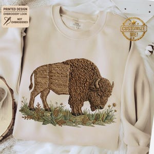 Op de afbeelding: Een beige sweatshirt met een gedetailleerd borduur-look ontwerp van een bizon. De bruine bizon staat op een stuk groen en bruin gras met kleine witte bloemen. De tekst "PRINTED DESIGN EMBROIDERY LOOK NOT EMBROIDERED" is zichtbaar.