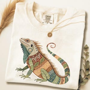Puede incluir: Camiseta color crema con una ilustración detallada de una iguana con intrincados patrones en tonos verdes, marrones y naranjas. La iguana está posada sobre un fondo blanco. La camiseta tiene una etiqueta Comfort Colors.