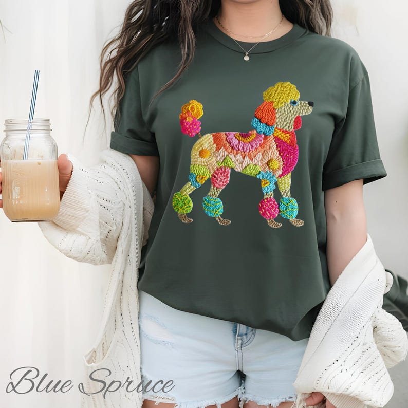 Pu&ograve; includere: T-shirt verde scuro con un colorato design di barboncino ricamato. Il barboncino &egrave; adornato con fiori e colori vivaci, tra cui giallo, rosa, arancione e blu. Il testo "Blue Spruce" &egrave; visibile in basso a sinistra.