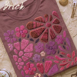 May include: A brick-coloured t-shirt featuring a large embroidered floral design. The embroidery consists of various flowers and petals in shades of pink, magenta, purple, and copper. The word Brick is printed in white script in the top left corner.