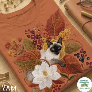 Könnte beinhalten: Ziegelrotes T-Shirt mit einer Stickerei einer Siamkatze mit blauen Augen, umgeben von Herbstblättern und Blumen in Orange-, Gelb- und Brauntönen. Unten befindet sich eine große weiße Blume. Der Text "YAM" befindet sich unten links.